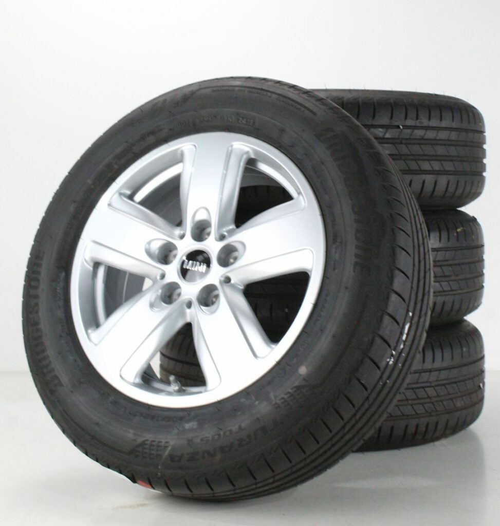 Four black Bridgestone car tires with silver alloy rims are stacked on a white surface. Three are stacked horizontally, while one stands upright at the front, displaying its five-spoke mini part rim and detailed tire tread pattern.